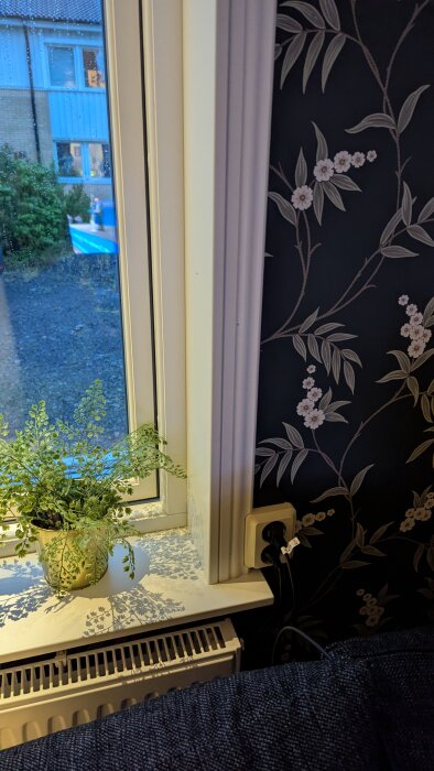 Fönster med bröstlist och blommig tapet, grön växt på fönsterbrädan, utsikt mot grannhus, element och vägguttag synliga.