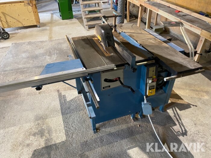 Justerbord sågmaskin i ett verkstadsutrymme med träbänk i bakgrunden.