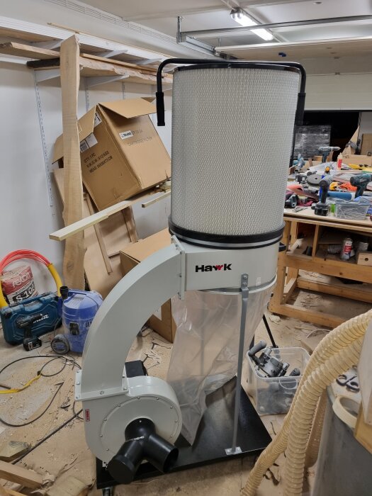 Dammsugare och spånhanteringsenhet i snickarverkstad, omgiven av verktyg, kartonger och sågspån.