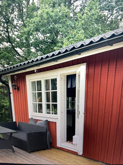 En röd fasad med vita fönster och dörr, ett svart möblemang på altanen, bredvid ett grönt skogsparti och en takränna ovanför.