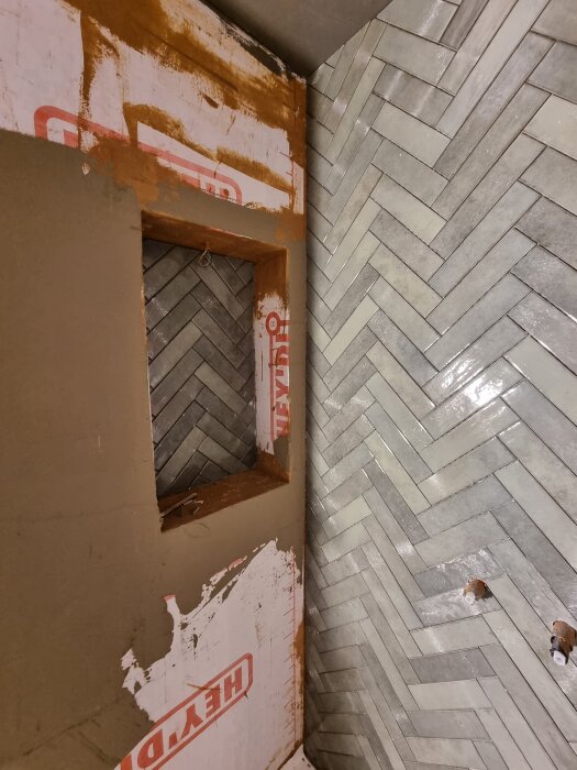 Duschnisch med fiskbensmönstrade kakelplattor och ofärdig konstruktion med byggmaterial.