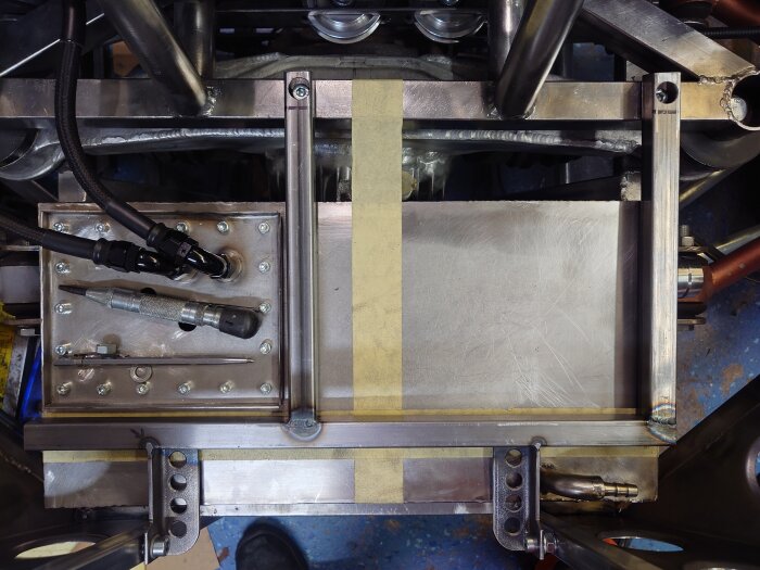 Metallram och monterad tank med röranslutningar i ett bilbygge, omgiven av förstärkta hållare och del av golvstruktur.