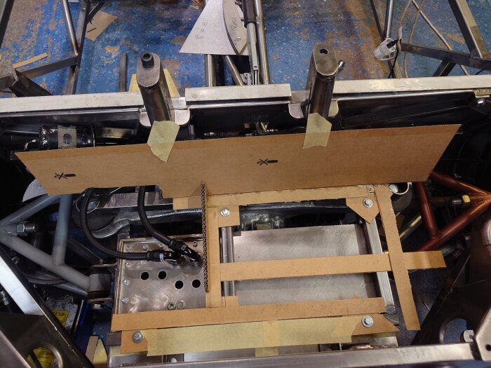 Kartongmallar skapade för montering av bränsletankfäste och golv i ett bagageutrymme, med metallram och slangar synliga.