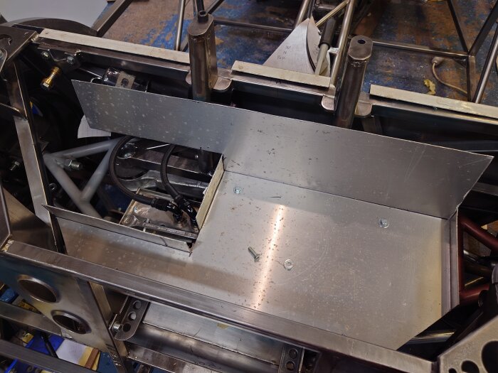 Metallkonstruktion i bagageutrymme, delvis monterat, med tankfäste och påbörjat golv. Rör och skruvar synliga i bakgrunden.