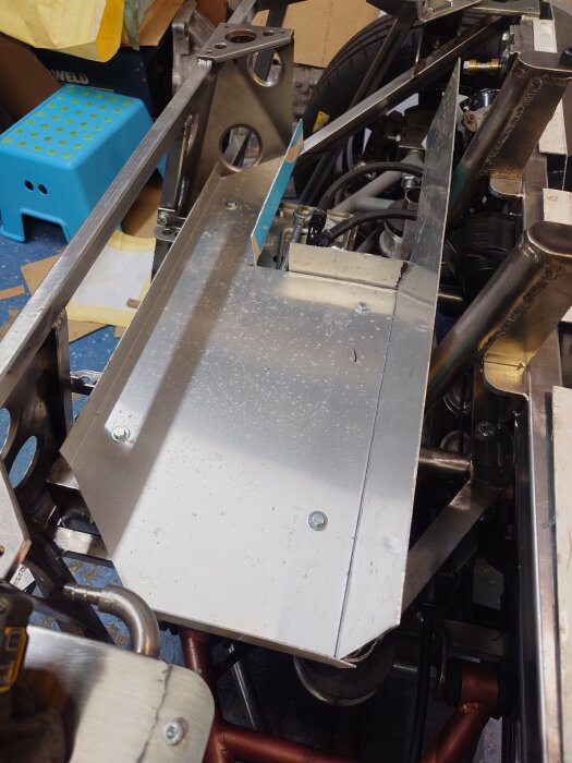 Metallplåt monterad i bilens bagageutrymme, omgiven av ramar och ledningar, med synlig bilhjul och verktyg i garage.