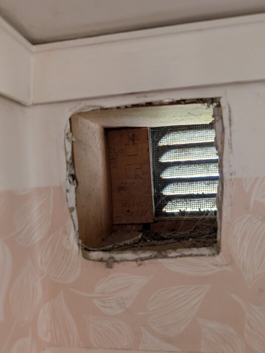 Rektangulärt ventilationshål i en vägg, med insyn till trälister och nät bakom, tapet med lövmönster runt hålet.