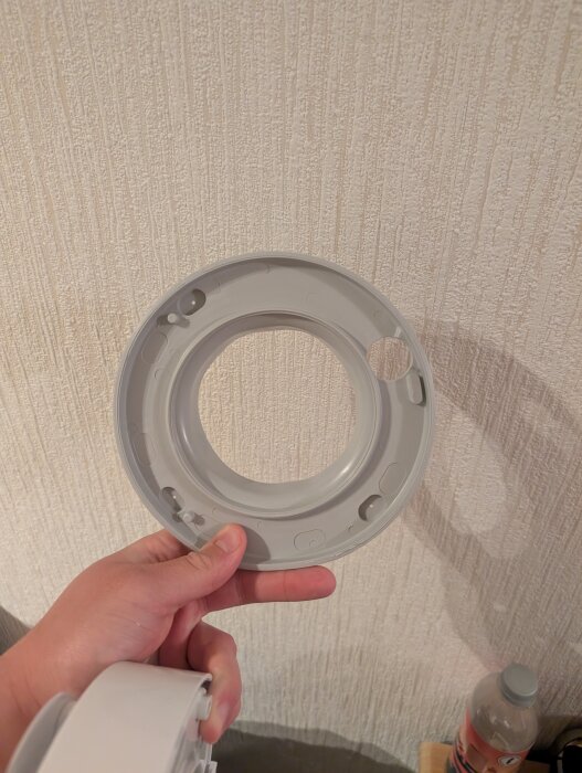 Hand som håller i en rund, grå plastdetalj mot en vit, texturerad vägg.