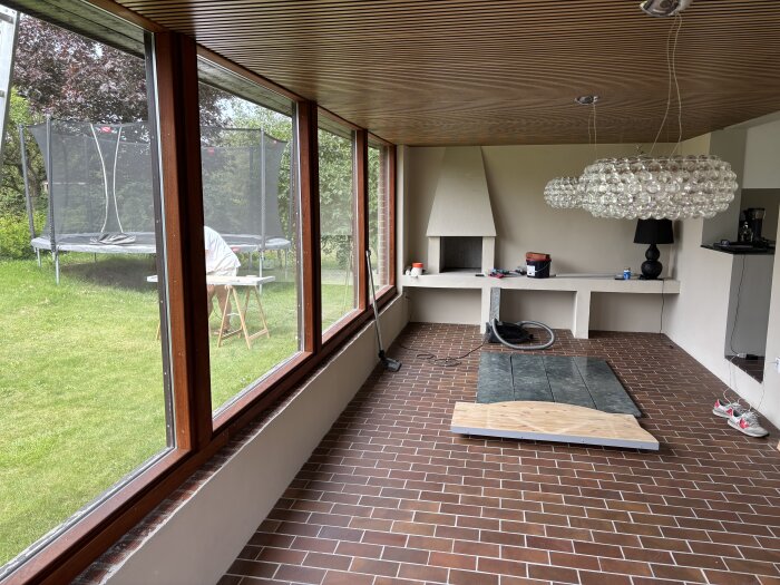 Interiör med stora fönster, en modern ljuskrona, braskamin och verktyg spridda på golvet. Utanför syns en trädgård med studsmatta.