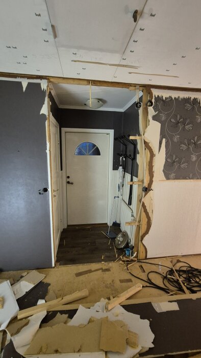 Rivningsarbete i hall, visar borttagen vägg med bröstpanel och exponerad dörröppning. Golvet är rörigt med byggmaterial och verktyg.