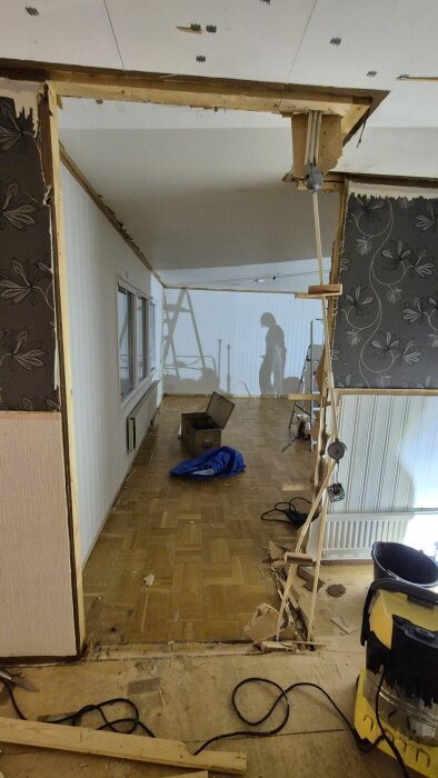 Renoveringsprojekt där en väggöppning skapats, exponerade ledningar och borttagen parkett syns. Skuggor av en person och verktyg i bakgrunden.