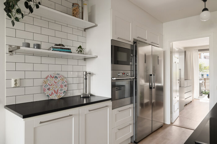 Modernt lantligt kök med vita skåpluckor, svart bänkskiva, vit kakelvägg och rostfria vitvaror. Öppna hyllor med böcker och dekoration.