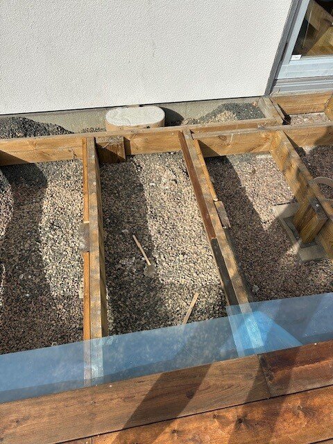 Trädäcksbygge med synliga reglar och grusunderlag, felaktigt placerade staketpinnar som stöd, saknade sågade ändar på trallen.