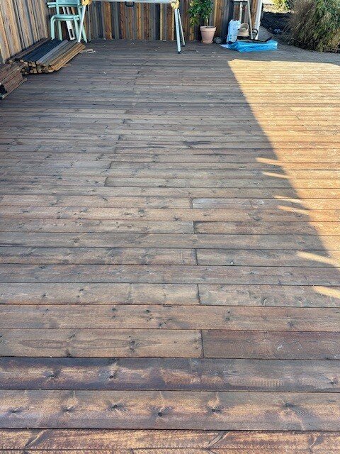 Trädäck med trätrall där skarvarna ej är rakt sågada och en del skarvar ligger i linje. Byggnadsdetaljer i bakgrunden.