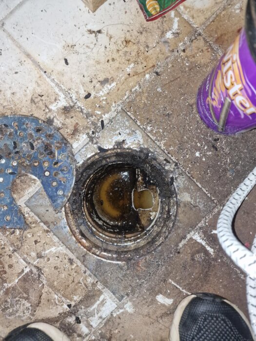 Badrumsavlopp med smuts och vatten runt, tecken på igensättning. En rengöringsprodukt synlig till höger.