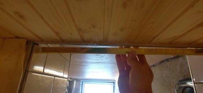 Hand som håller mässingslist mot träram runt bastuingång, tak i trä och kakel synliga i bakgrunden.