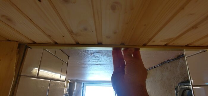 Hand håller mässingslist vid bastuingångens tak i trä, med spegel och vägg i bakgrunden.