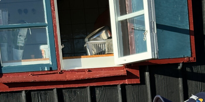 Öppet fönster med blå fönsterluckor och inredning inuti, inklusive kastruller på en diskställ i ett hus nära stranden.