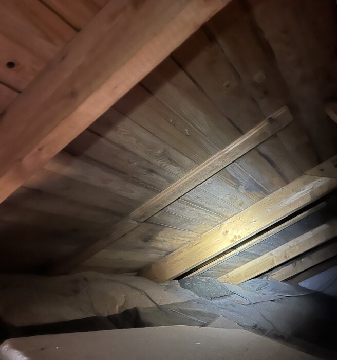Träbjälkar och takpanel i ett vindsutrymme, med isolering synlig i hörnet, belyst av ficklampa.