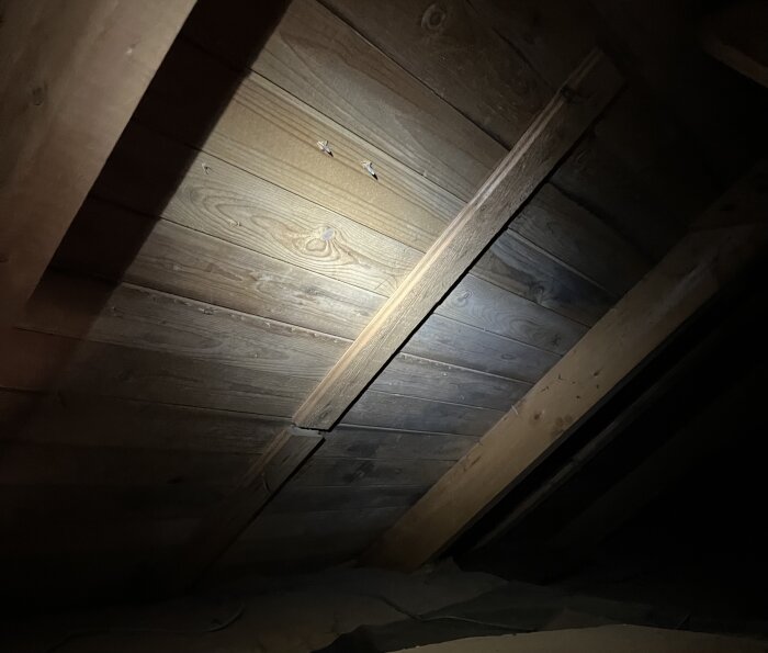 Träpanel i ett tak fotograferat i dåligt ljus, fokus på brädor och stöd under renoveringsinspektion för att kontrollera mögel eller röta.