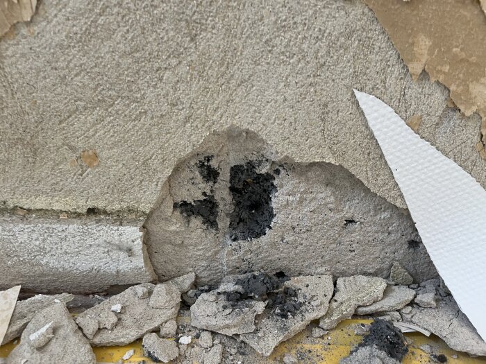 Närbild av en vägg med skadad puts och exponerad blåbetong efter en reva som visar svart missfärgning och sönderfallna bitar på golvet.