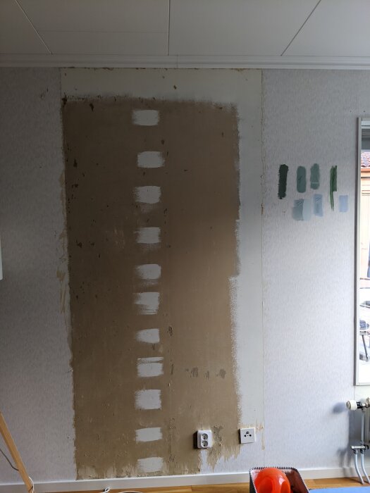 Delvis borttagen tapet på en vägg med synliga gipsskivor. Prover med olika färger målade på väggen till höger.
