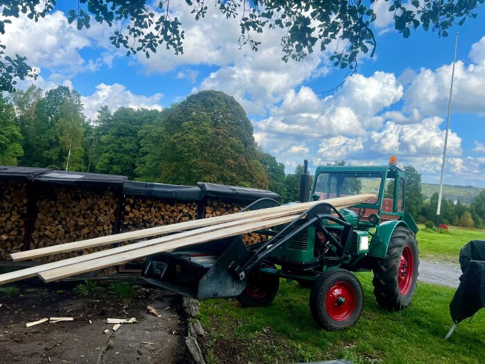 Traktor lastar brädor på en gräsmatta med vedstapel i bakgrunden, under en klarblå himmel med moln.