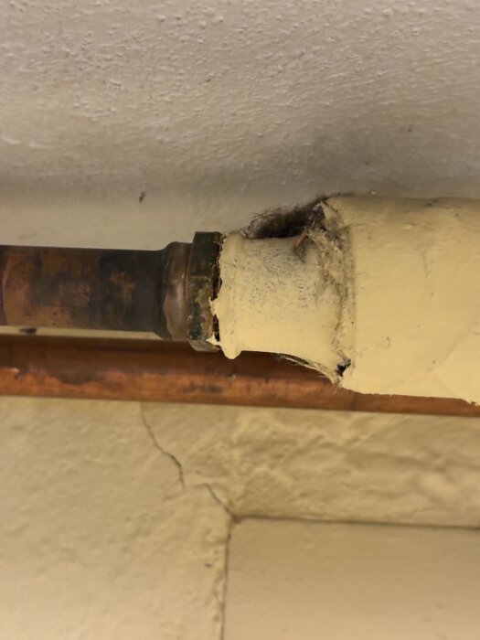 Rostigt rör med skadad gul isolering och synliga fibrer i källare, möjligen asbest, beläget mot sprucken vägg.