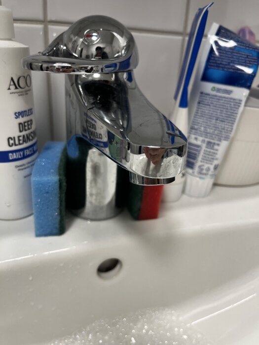 Tvättblandare med kromfinish monterad på ett handfat, omgiven av rengöringsprodukter och svampar.