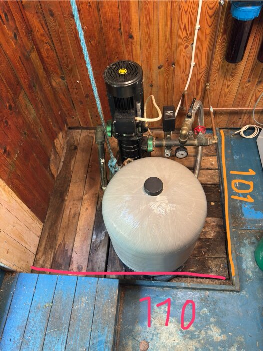 Vattenpump och tank i träkällare med märkning av mått i rött och orange, blå golv och väggar, samt synliga rör och filter.