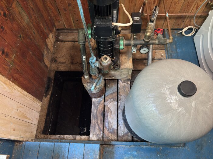 Brunn med vattenpump i källare, delvis vattenfylld, trälåda sänkt i vattnet, och betong på bakre delen. Rustik omgivning med träpanel.