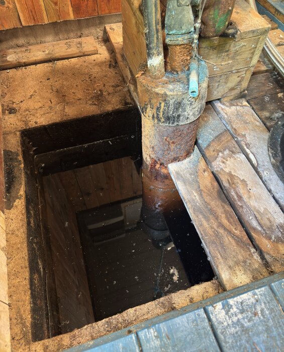 Rostig vattenpump och brunn i träkällare, delvis vattenfylld.