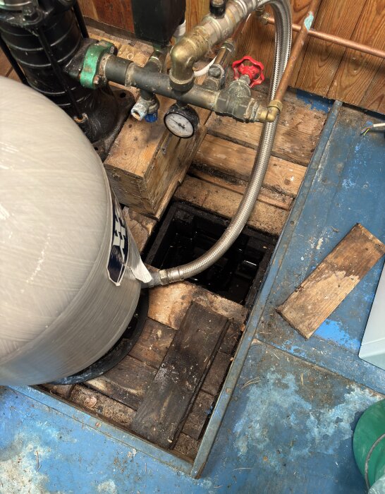 Vattenpump och ledningar vid en brunn i källare. Pumpen är monterad på träplankor över en öppen brunnyta med metalldelar och vattenledningar.