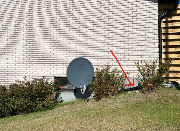 Fasad med satellitdisk och buskar där en platonmatta kan skymtas bakom buskarna, markerad med en röd pil.