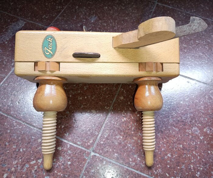 Äldre träbaserad hyvel med gängade delar av trä och metallhyvelstål, stående på rödbrunt tegelgolv.