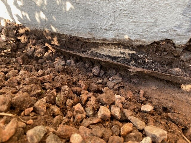 Grus vid husvägg med presenning och betongvägg i källaren synlig, delvis rengjort område.