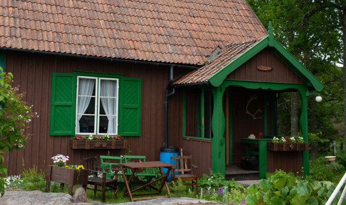 Gammalt sommarhus från 1926 med gröna luckor på nedervåningen, verandan och ett blomsterprytt fönster mot en lummig bakgrund.