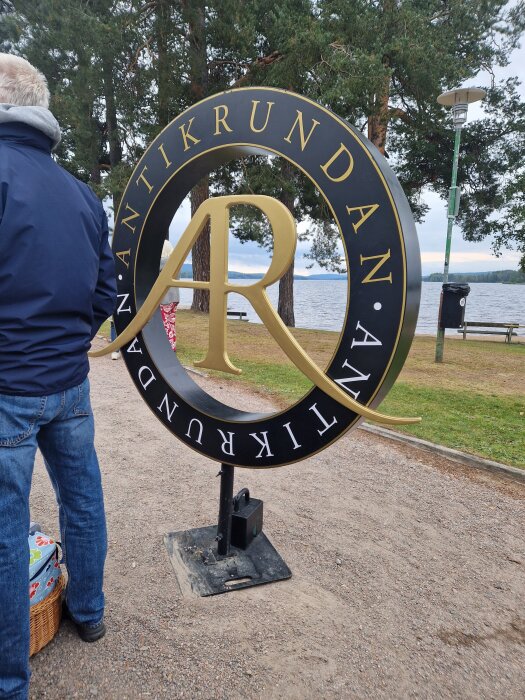 Skylt för "Antikrundan" vid en sjö med träd och parkbänkar i bakgrunden. En person står bredvid skylten med en picknickkorg.