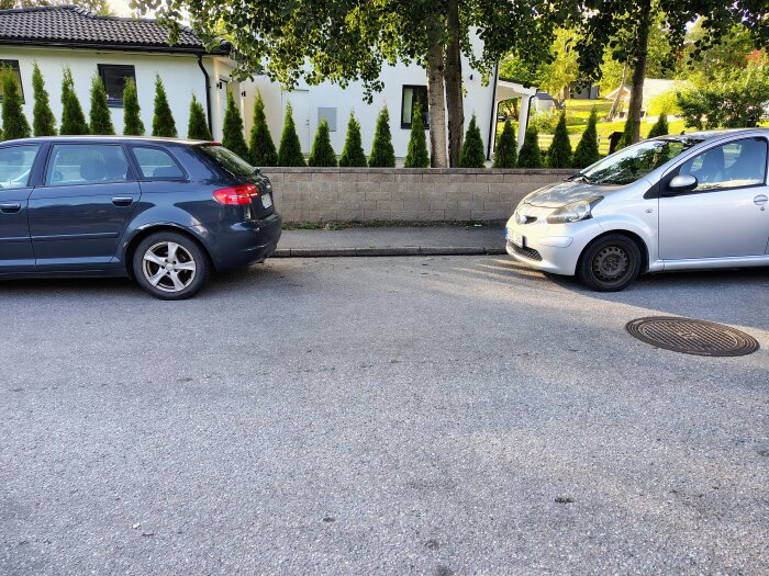 Mörk och silverfärgad bil parkerade nära varandra på gata, nästan utan mellanrum.