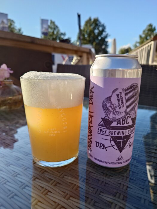 Burk och glas med öl från Apex Brewing Company, etiketten visar "Duskwatch DIPA" mot en bakgrund av en solig uteservering.
