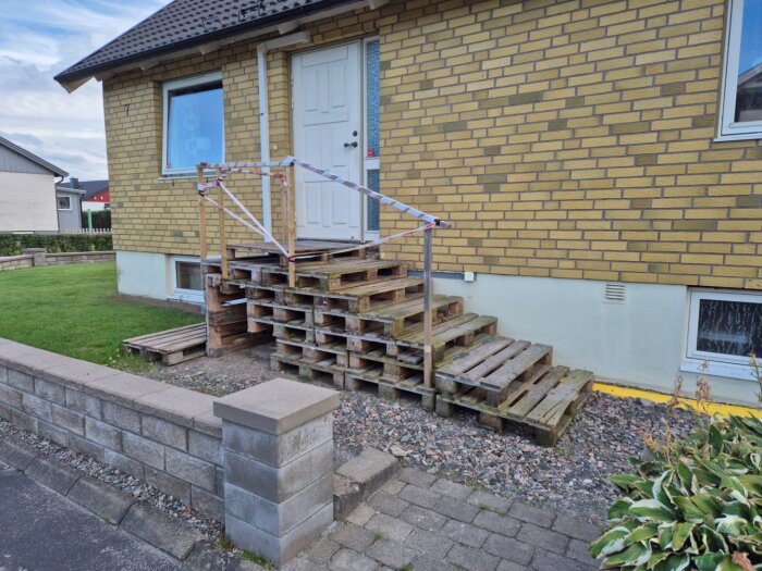 Trappkonstruktion av lastpallar leder upp till en dörr på ett tegelhus. En temporär räcke av trä och tejp är fäst vid trappan.