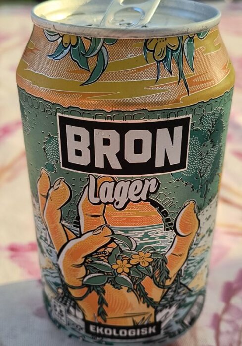 Burk med Bron Lager, ekologisk öl med färgglad illustrerad design som visar en hand och växter framför en solnedgång över vatten.