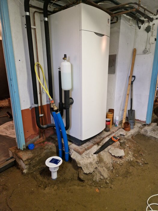 Bergvärmepump placerad på plattform av tegel och betong i källare, omgiven av rör och verktyg, med murad vägg i bakgrunden.