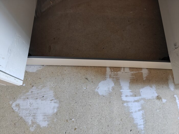 Öppet utrymme under köksskåp med synligt golv och vit sockel framtill. Golvet visar färgfläckar och ytan ser något grov ut.