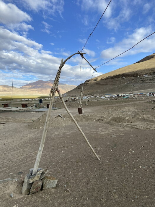 Provisorisk elledning i Himalaya, uppställd med trästolpar och stenar, under blå himmel med berg och by i bakgrunden.