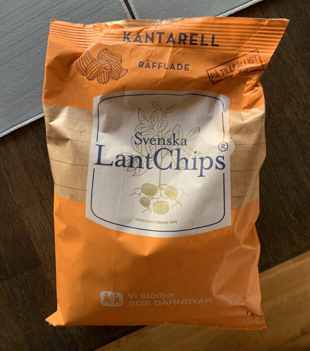 En påse svenska LantChips med kantarellsmak, räfflade, på ett bord. Säsongsprodukt, text "Vi stödjer SOS Barnbyar" synlig.