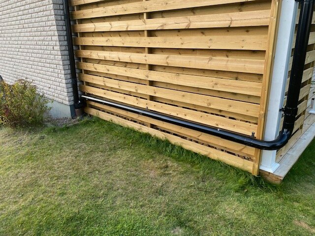 Svart stuprör anslutet vid hörnet av en träpanel intill en tegelvägg, över gräs.