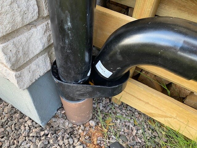 Svart stuprör anslutet till krökt rör vid hörnet av en byggnad, omgiven av grus och träpaneler.