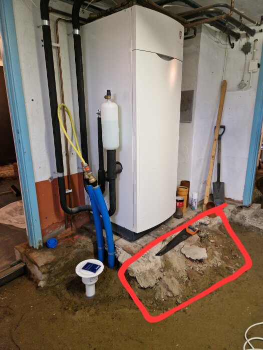 Värmepump i källare med rör och verktyg, bitar av betong och murbruk på marken framför, markerat område med såg på toppen.