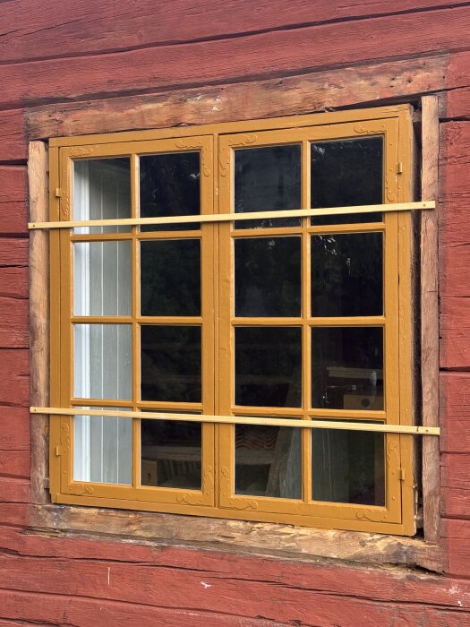 Gult fönster på röd träfasad, delvis renoverat med nya trädetaljer, visar fönsterrenovering med detaljerade träornament.