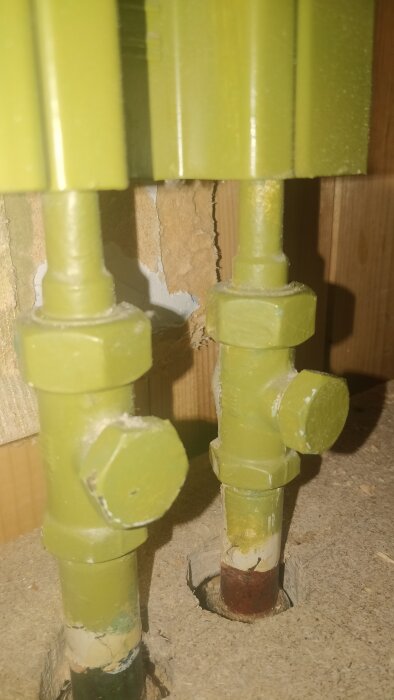 Närbild av två gröna rörkopplingar till radiator med ventiler, infällda i golvet. Bakgrunden visar gipsvägg och träpanel.